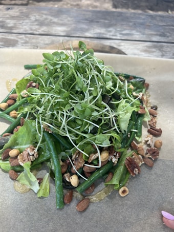 A fresh salad composed of leafy greens, green beans, and a variety of nuts such as almonds and walnuts is arranged on a sheet of parchment paper. The mixture is topped with numerous sprouts, creating a vibrant and rustic display.
