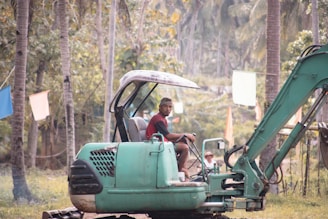A man in a red shirt and cap operates a green excavator surrounded by a tropical landscape with palm trees and colorful flags hanging in the background.
