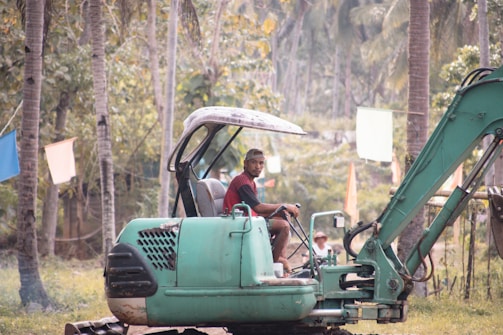 A man in a red shirt and cap operates a green excavator surrounded by a tropical landscape with palm trees and colorful flags hanging in the background.