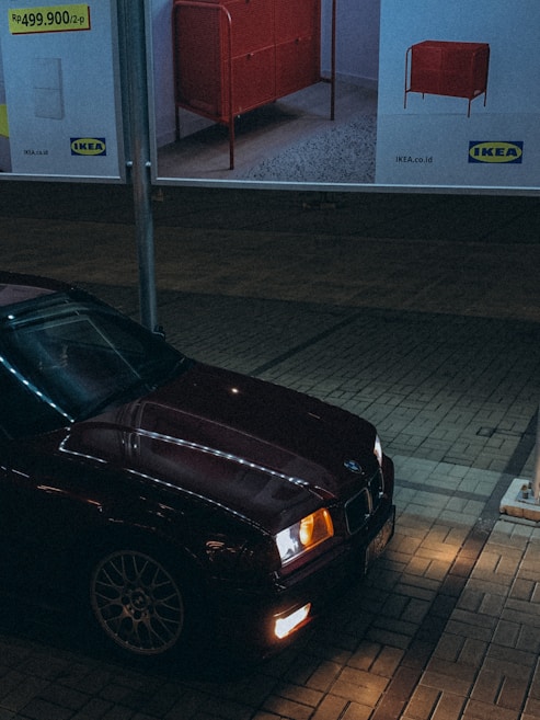 A dark city scene featuring a red car parked under a billboard for IKEA furniture. The car's headlights are on, illuminating the brick pavement. The billboard above displays an advertisement for a red piece of furniture with the IKEA logo and pricing details in the local currency.