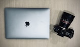A silver laptop with an Apple logo is placed on a wooden surface next to a black Sony camera with a large lens.
