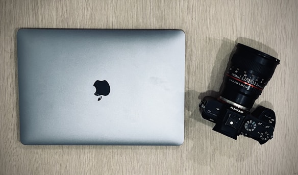 A silver laptop with an Apple logo is placed on a wooden surface next to a black Sony camera with a large lens.