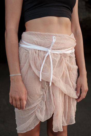 A person wearing a black cropped top and a light pink sheer skirt tied with a white ribbon. The fabric has a soft, flowing texture, and the skirt is layered, providing an elegant appearance.