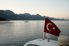A scenic view of calm water with a boat in the background, surrounded by distant mountains under a clear sky. A Turkish flag is prominently displayed on a boat in the foreground, fluttering gently.