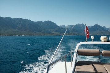A small boat is moving across a calm blue sea with a distant view of mountainous landforms under a clear sky. The rear of the boat features a seating area and a national flag, suggesting a leisurely cruise. The boat's wake creates a gentle pattern on the water's surface.