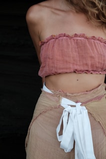 A person is wearing a pink bandeau-style top and a beige wrap skirt with a white ribbon or belt tied around the waist. The image captures the upper body from the shoulders to just below the waist. The background is dark and contrasts with the clothing.