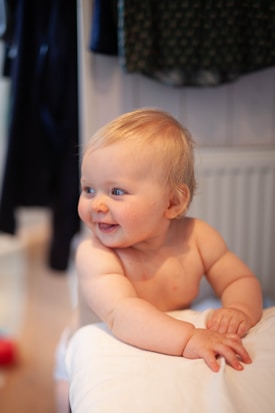 A smiling baby with light blonde hair gazes to the side while leaning on a white surface. The baby is shirtless, showing fair skin with a slight reddish tone on the cheeks. The background is blurred, featuring some dark-colored garments hanging and part of a radiator.