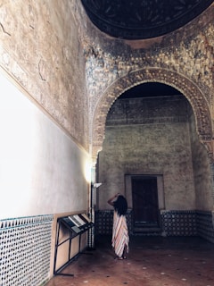 An intricately designed interior features ornate arches and wall carvings. A person in a colorful, striped dress stands with their back to the viewer, taking a photo with a phone. The room has a historical or cultural aesthetic, with decorative tiles and dim lighting that accentuates the architectural details.