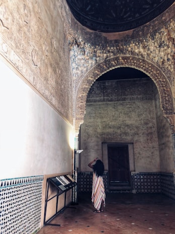 An intricately designed interior features ornate arches and wall carvings. A person in a colorful, striped dress stands with their back to the viewer, taking a photo with a phone. The room has a historical or cultural aesthetic, with decorative tiles and dim lighting that accentuates the architectural details.