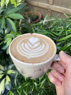 Close-up of a hand holding a nude beige ceramic cup filled with frothy cappuccino, set against a light lavender background.