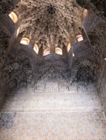 An intricately designed interior with detailed geometric and floral motifs on the walls and ceiling. The architecture features a series of small, arched windows that allow natural light to illuminate the space, highlighting its ornate plaster work.