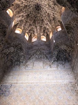 An intricately designed interior with detailed geometric and floral motifs on the walls and ceiling. The architecture features a series of small, arched windows that allow natural light to illuminate the space, highlighting its ornate plaster work.
