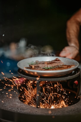 A close-up of a frying pan on a stove, sizzling with what appears to be steak garnished with herbs. Sparks fly around, suggesting a high heat source, possibly from a wood or charcoal fire. The background is dark, creating a dramatic contrast with the bright sparks and the cooked food.
