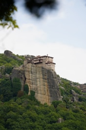 A large rock formation hosts a historic monastery perched at its peak. The structure is built of stone and blends seamlessly with the natural landscape. Lush green trees surround the base, and the sky is overcast with a hint of blue.