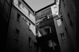 A black and white photograph of an interior courtyard surrounded by multi-story residential buildings. The image captures the play of light and shadow across the walls and windows. Balconies with potted plants and air conditioning units are visible. The architectural style appears old, with narrow and tall walls creating a sense of enclosure.