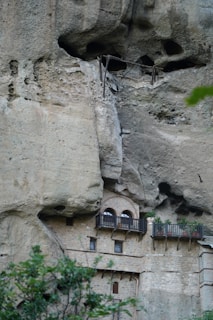A stone building is integrated into a high cliff, featuring several arched windows and balconies with railings and potted plants. The cliff face shows natural erosion patterns and cavities above the structure.