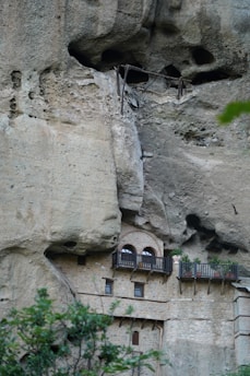 A stone building is integrated into a high cliff, featuring several arched windows and balconies with railings and potted plants. The cliff face shows natural erosion patterns and cavities above the structure.