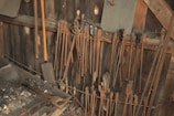 A collection of blacksmith tools hangs on a wooden wall in a workshop setting. Long tongs are lined up neatly, alongside a couple of wooden-handled shovels. The surface below them is covered in ash and coal remnants, suggesting recent use.