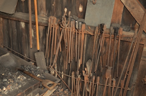 A collection of blacksmith tools hangs on a wooden wall in a workshop setting. Long tongs are lined up neatly, alongside a couple of wooden-handled shovels. The surface below them is covered in ash and coal remnants, suggesting recent use.