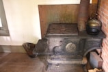 An antique cast iron stove with ornate detailing, placed against a wall. The stove top features four circular plates, and there is a metal kettle resting on it. The surrounding area includes a brick wall on one side and a bucket on the wooden floor beside the stove.