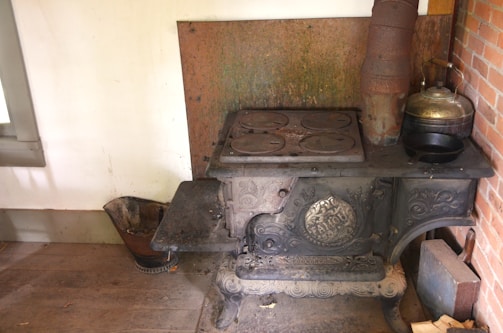 An antique cast iron stove with ornate detailing, placed against a wall. The stove top features four circular plates, and there is a metal kettle resting on it. The surrounding area includes a brick wall on one side and a bucket on the wooden floor beside the stove.
