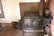 An antique cast iron stove with ornate detailing, placed against a wall. The stove top features four circular plates, and there is a metal kettle resting on it. The surrounding area includes a brick wall on one side and a bucket on the wooden floor beside the stove.