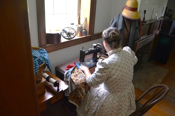 A woman in a floral dress is seated at a vintage sewing machine, working on fabric with different patterns. The setting appears to be an old-fashioned room with wooden furniture, including a table holding spools of thread and fabrics. A large window allows natural light to enter, highlighting the sewing area. Beside her, a mannequin wearing a straw hat and a coat stands near a fireplace.
