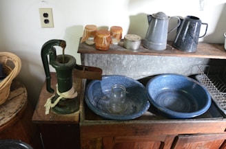 A rustic kitchen setting features a vintage hand pump on an old wooden counter next to blue enamel bowls and a clear glass jar. There are two metal pitchers and jars with preserves on a shelf above. The scene includes a basket and various kitchen accessories, suggesting a traditional and functional environment.