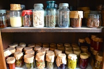 Shelves filled with various jars, some sealed with paper and string, containing preserved foods such as vegetables, beans, and spices. The jars are made of glass and vary in size and shape, containing an assortment of natural ingredients.