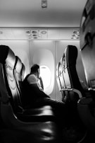 A person is seated on a plane, looking out of the window. The interior of the aircraft, including the overhead compartments and seats, is visible. The lighting suggests it's a daytime flight, with bright light coming through the window.