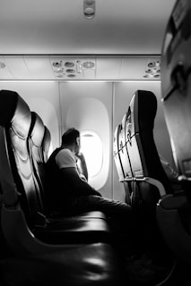 A person is seated on a plane, looking out of the window. The interior of the aircraft, including the overhead compartments and seats, is visible. The lighting suggests it's a daytime flight, with bright light coming through the window.