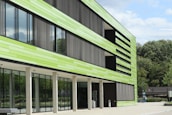 A modern building with a facade made of horizontal green panels and large glass windows. The building has vertical slats that appear to function as sunshades. It is situated in a landscaped area with trees in the background and a clear blue sky.