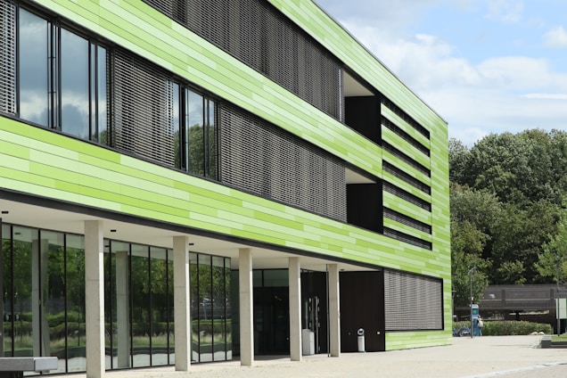 A modern building with a facade made of horizontal green panels and large glass windows. The building has vertical slats that appear to function as sunshades. It is situated in a landscaped area with trees in the background and a clear blue sky.