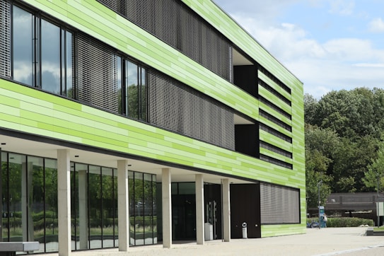 A modern building with a facade made of horizontal green panels and large glass windows. The building has vertical slats that appear to function as sunshades. It is situated in a landscaped area with trees in the background and a clear blue sky.