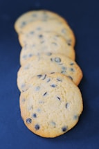 Five chocolate chip cookies are arranged in a slightly overlapping line, set against a dark blue background. The cookies have a golden-brown color with visible chocolate chips embedded in them, indicating they are freshly baked.