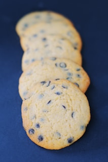 Five chocolate chip cookies are arranged in a slightly overlapping line, set against a dark blue background. The cookies have a golden-brown color with visible chocolate chips embedded in them, indicating they are freshly baked.
