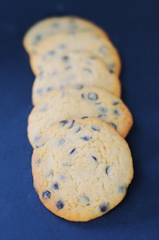 Five chocolate chip cookies are arranged in a slightly overlapping line, set against a dark blue background. The cookies have a golden-brown color with visible chocolate chips embedded in them, indicating they are freshly baked.