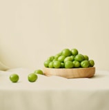 A wooden bowl is filled with green plums, resting on a light-colored fabric surface. A few plums are scattered outside the bowl, creating a natural and simple composition.