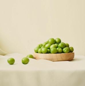 A wooden bowl is filled with green plums, resting on a light-colored fabric surface. A few plums are scattered outside the bowl, creating a natural and simple composition.