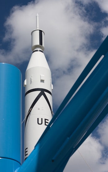 A white rocket with black markings is pointing towards the sky, framed against a backdrop of scattered clouds. In the foreground, there are large blue metallic structures, possibly part of a launch pad or support system.