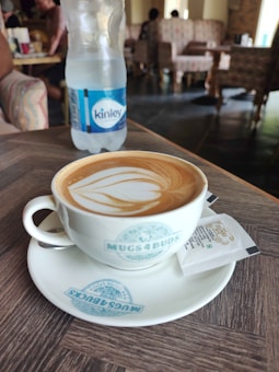 A coffee cup filled with a latte art design sits on a branded saucer on a wooden table. Beside it is a small white packet. In the background, there is a plastic water bottle labeled 'Kinley'. The setting appears to be a cozy cafe with upholstered chairs and soft lighting.