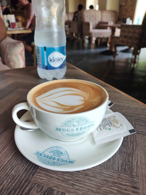 A coffee cup filled with a latte art design sits on a branded saucer on a wooden table. Beside it is a small white packet. In the background, there is a plastic water bottle labeled 'Kinley'. The setting appears to be a cozy cafe with upholstered chairs and soft lighting.