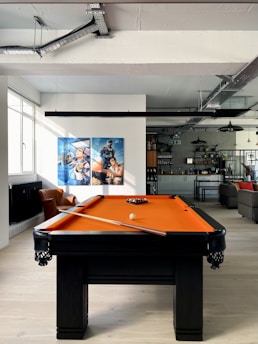 A modern, well-lit room featuring an orange pool table with cues resting on it. In the background, there is an open kitchen with industrial-style decor, visible pipes, and ceiling beams. Two framed artworks, depicting astronauts, are mounted on the wall above a brown armchair. Natural light streams in from a large window to the left.