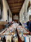 A large room with high arched ceilings contains numerous tables filled with books, suggesting a book sale or library event. The architecture features tall, arched windows and a prominent organ at the back. Several people are browsing the books, creating a lively atmosphere.