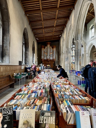 A large room with high arched ceilings contains numerous tables filled with books, suggesting a book sale or library event. The architecture features tall, arched windows and a prominent organ at the back. Several people are browsing the books, creating a lively atmosphere.