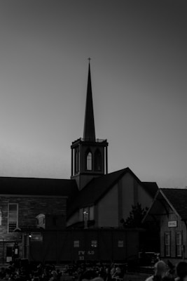 A black and white photograph of a church with a tall, pointed steeple. The building is silhouetted against the twilight sky, with detailed architectural features visible on the facade. Below the church, there is a crowd gathered, possibly attending an event.
