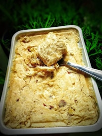 A rectangular dish filled with a textured, fibrous, beige-colored sweet, with pieces of nuts embedded within it. A metal spoon is placed on top of the sweet, scooping out a chunk. The background consists of green foliage, providing a natural contrast to the dish.