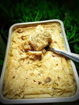 A rectangular dish filled with a textured, fibrous, beige-colored sweet, with pieces of nuts embedded within it. A metal spoon is placed on top of the sweet, scooping out a chunk. The background consists of green foliage, providing a natural contrast to the dish.