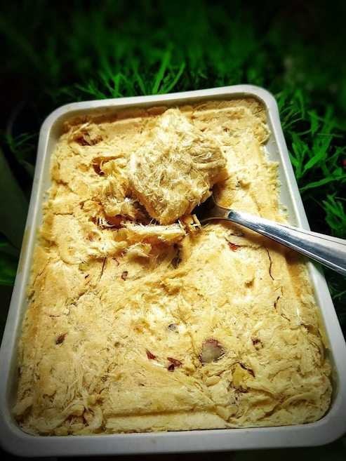 A rectangular dish filled with a textured, fibrous, beige-colored sweet, with pieces of nuts embedded within it. A metal spoon is placed on top of the sweet, scooping out a chunk. The background consists of green foliage, providing a natural contrast to the dish.