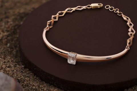 A delicate gold bracelet with an intricate chain design and a central rectangular diamond set in a minimalist and elegant manner. The bracelet is placed on a dark circular platform against a textured sandy surface, showcasing its luxurious and refined craftsmanship.